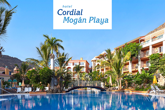 Blick über Hotelanlage mit roten Gebäuden, unten am Bild und in der Mitte Pools zu sehen, am Horizont eine Gebirgskette